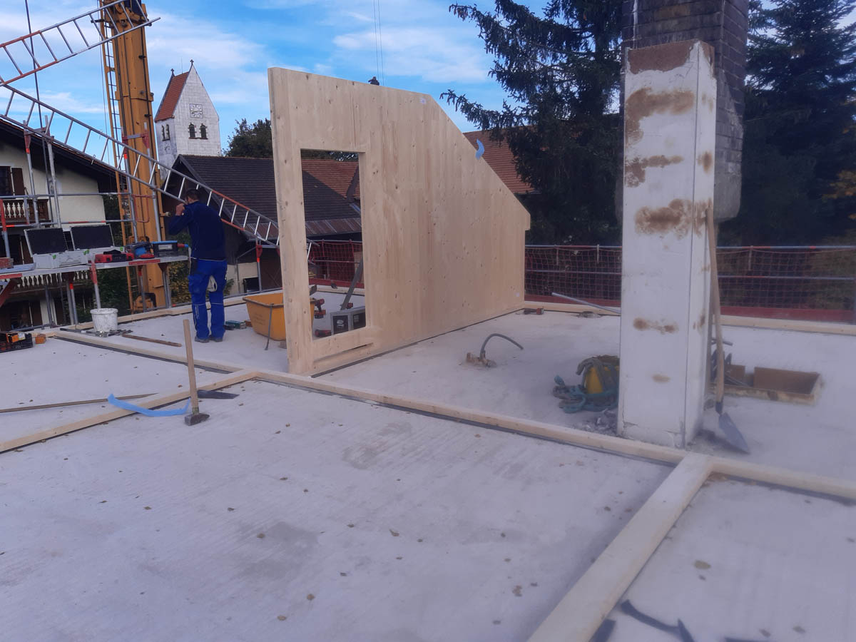 Baustelle mit einem Arbeiter, der eine Leiter hebt, und einer teilweise montierten Holzwand auf einem Betonboden.