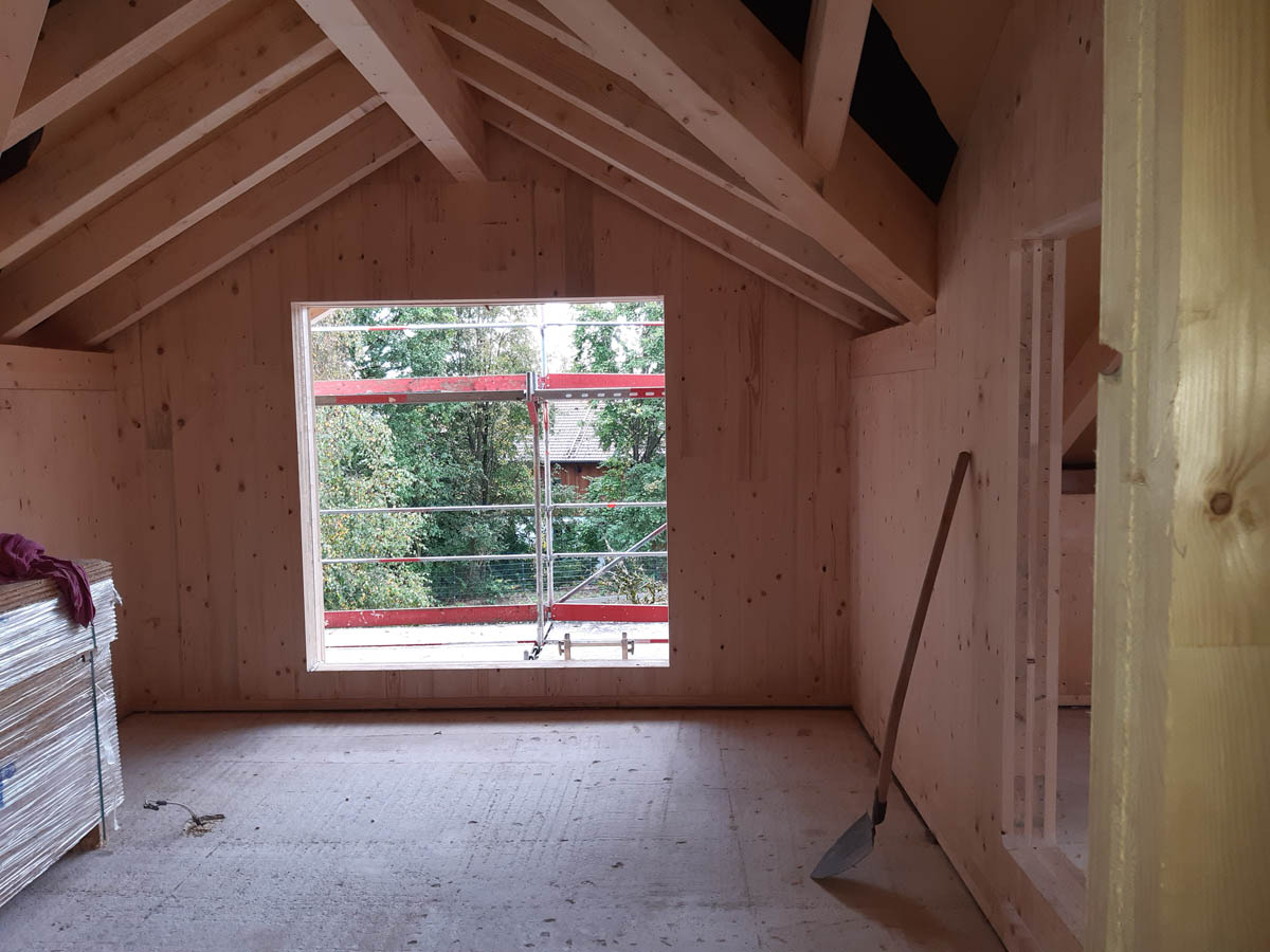 Innenraum eines Rohbaus mit Holzwänden und Dachstuhl, offenem Fensterrahmen mit Blick auf Bäume und Gerüst.