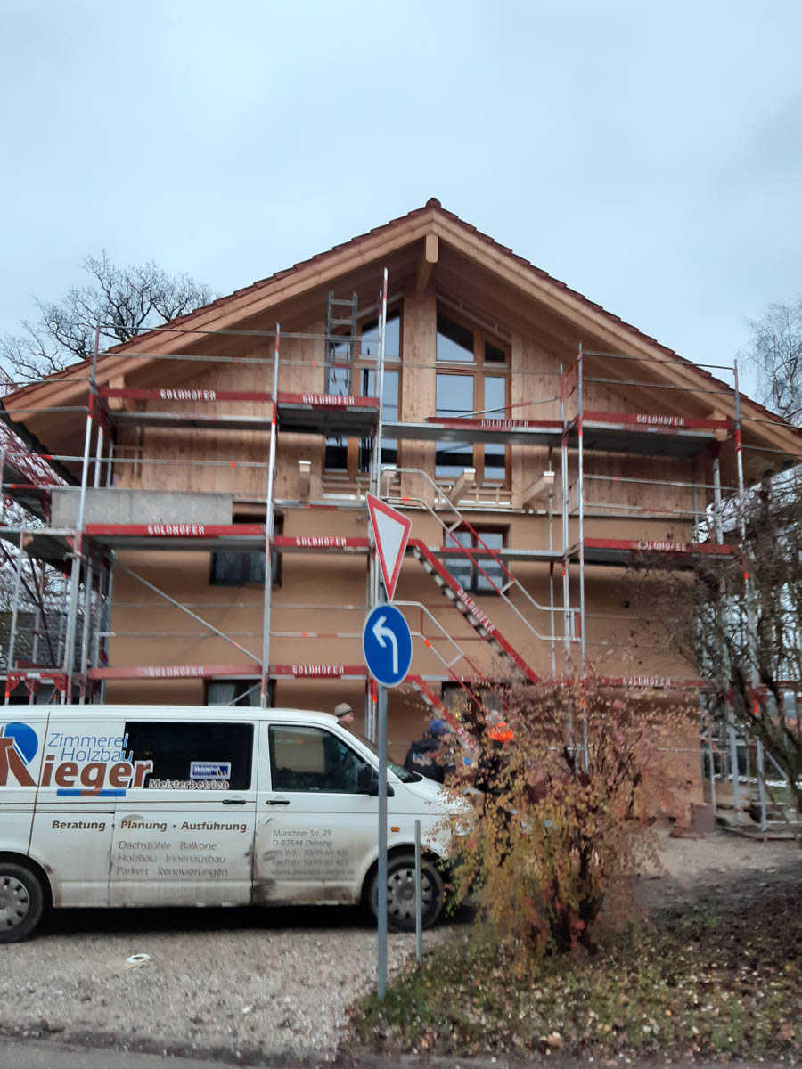 Ein mehrstöckiges Wohnhaus mit Holzfassade im Rohbau, umgeben von Gerüsten, davor ein weißer Handwerks-Van und ein Verkehrsschild für Linksabbiegen.
