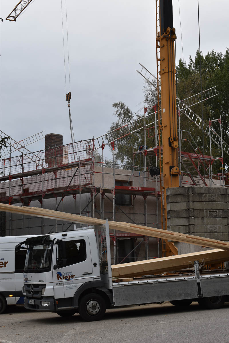 Lastwagen mit Holzbaumaterial vor einer Baustelle mit Gerüst und Baukran unter grauem Himmel.
