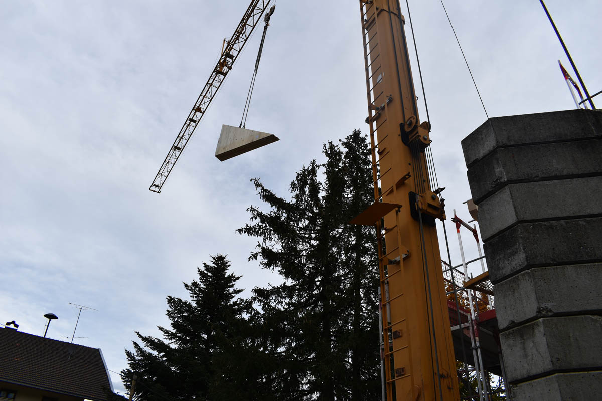 Ein Kran hebt ein dreieckiges Betonelement auf einer Baustelle neben Tannenbäumen und Gebäuden.