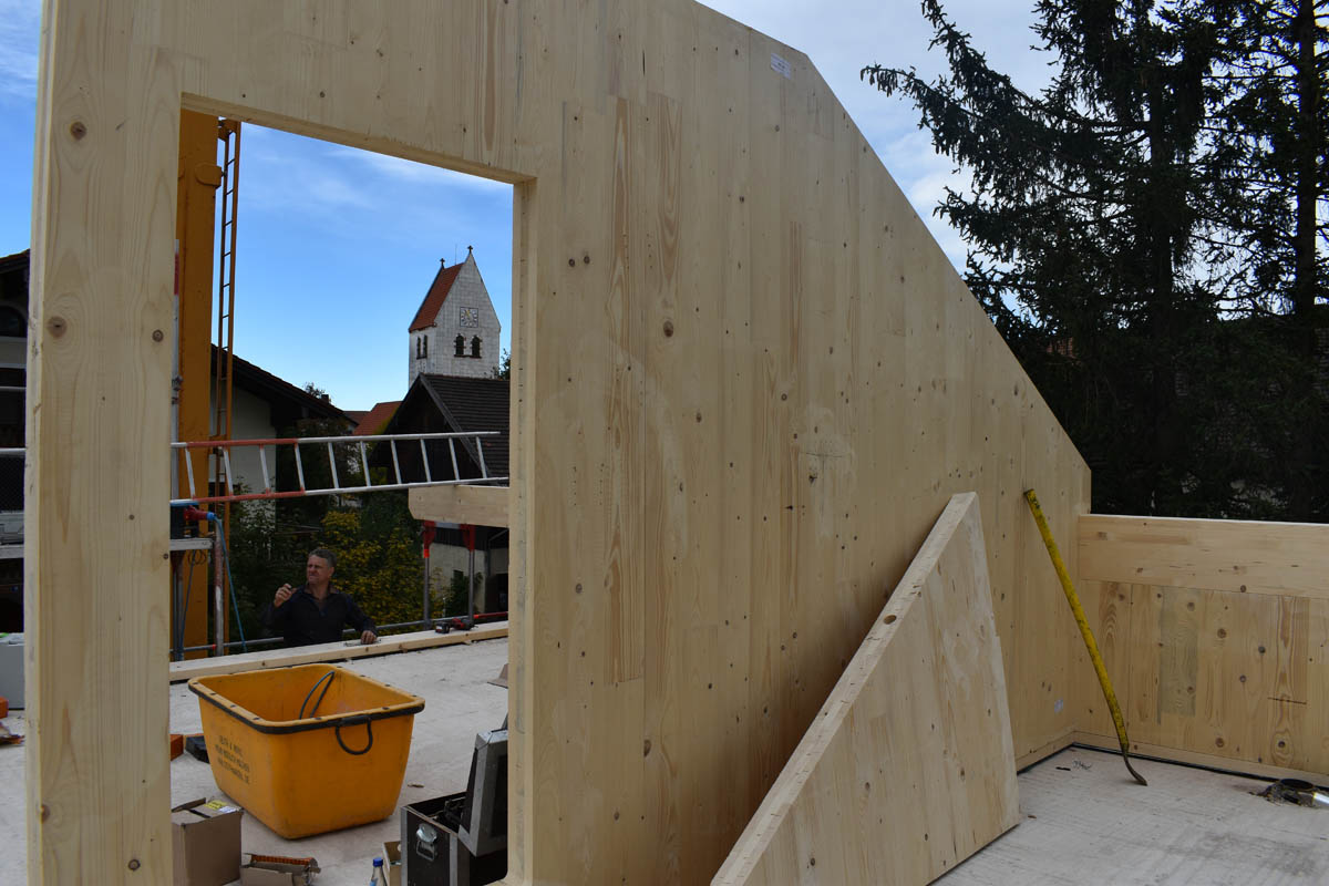 Bauarbeiter auf einer Baustelle mit großen Holzpaneelen, gelbem Behälter und im Hintergrund Kirchturm.
