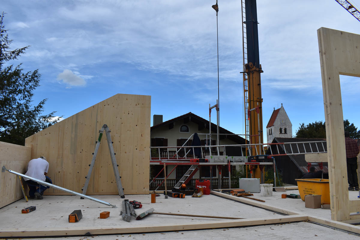 Baustelle mit Holzrahmenwänden, Werkzeugen auf Boden und einem Kran vor blauem Himmel in einer Wohngegend.