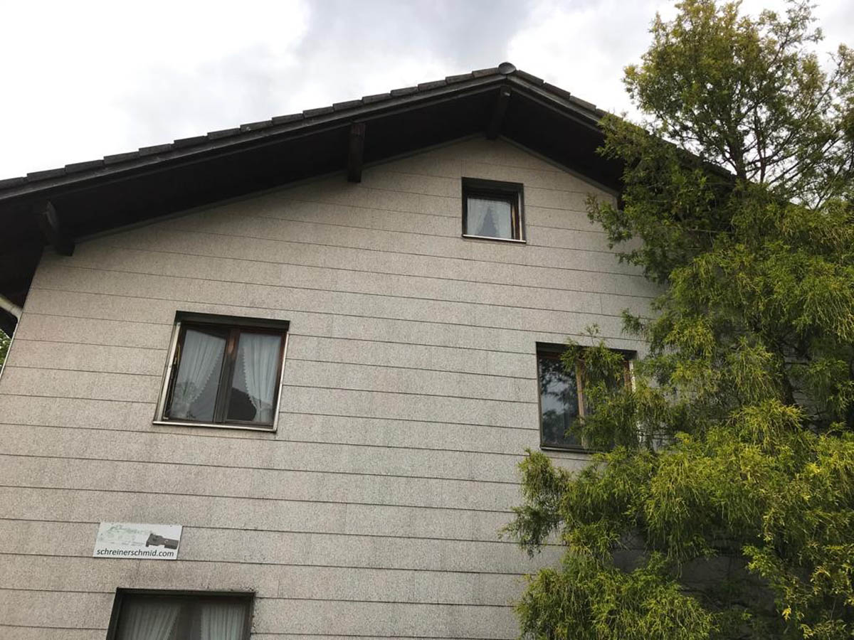 Seitliche Ansicht eines grauen Hauses mit drei Fenstern und einem Baum rechts.