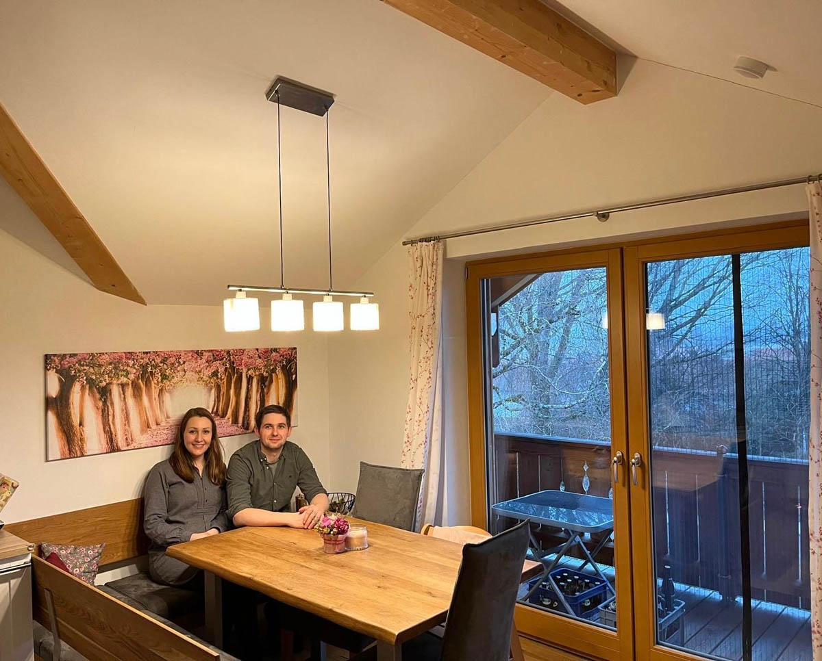 Ein Mann und eine Frau sitzen an einem Esstisch in einem gemütlichen Raum mit Holzbalken und großer Glasschiebetür zum Balkon.