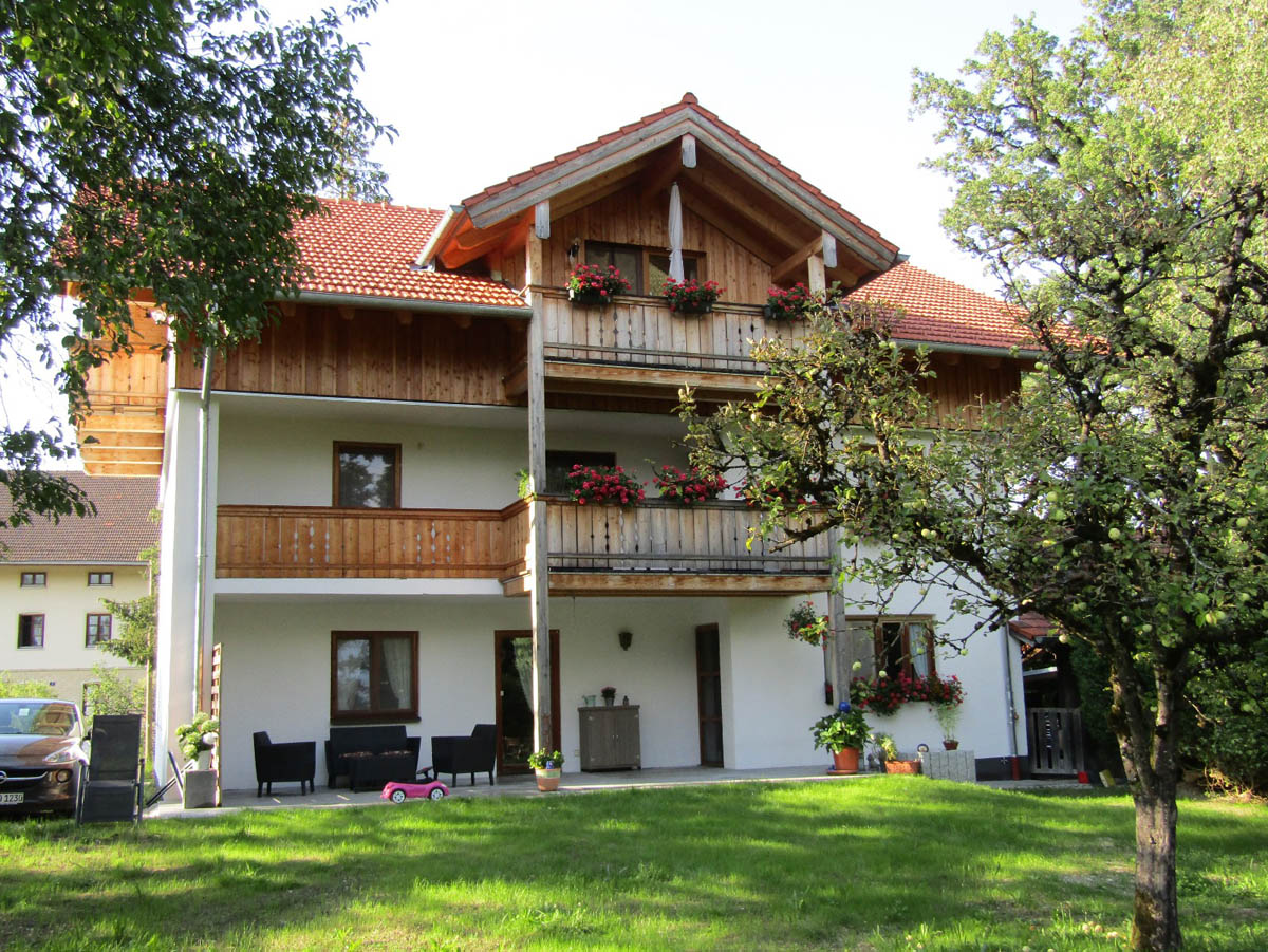 Dreistöckiges Haus mit Holzverkleidung, roten Blüten auf Balkonen und grünem Garten mit Baum im Vordergrund.