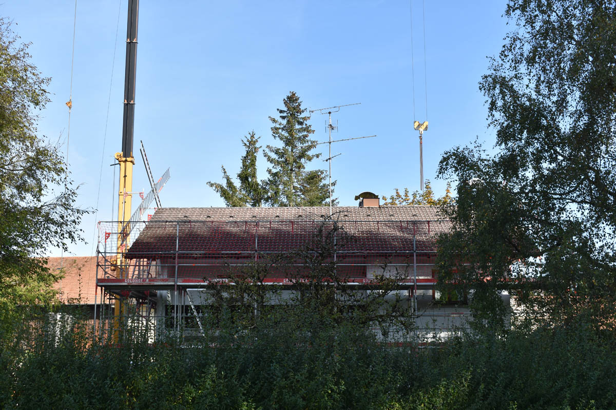 Haus mit Ziegeldach und Baugerüst, umgeben von Bäumen und Sträuchern bei klarem Himmel.