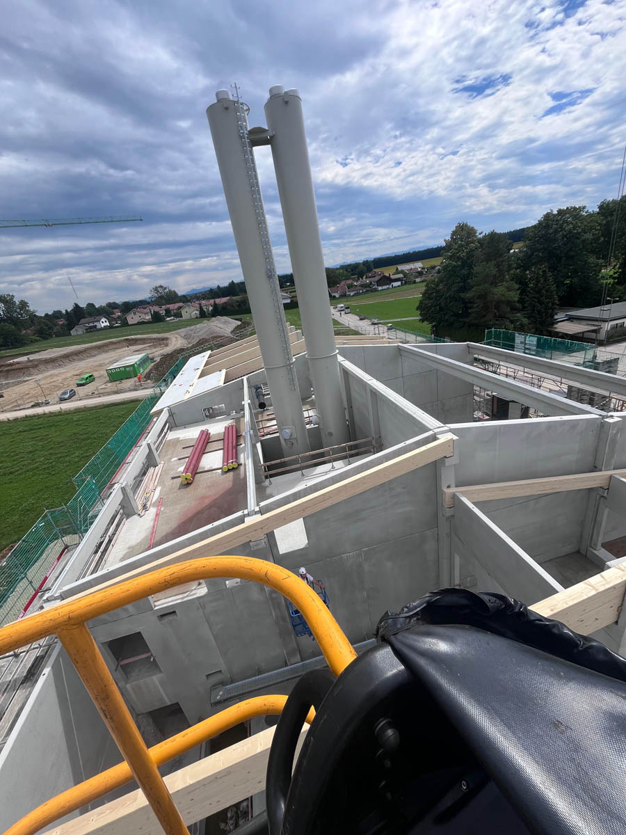 Blick von einer gelben Geländerplattform auf eine Baustelle mit hohen zwei weißen Industrieabgasrohren und teilweise errichteten Betonwänden.