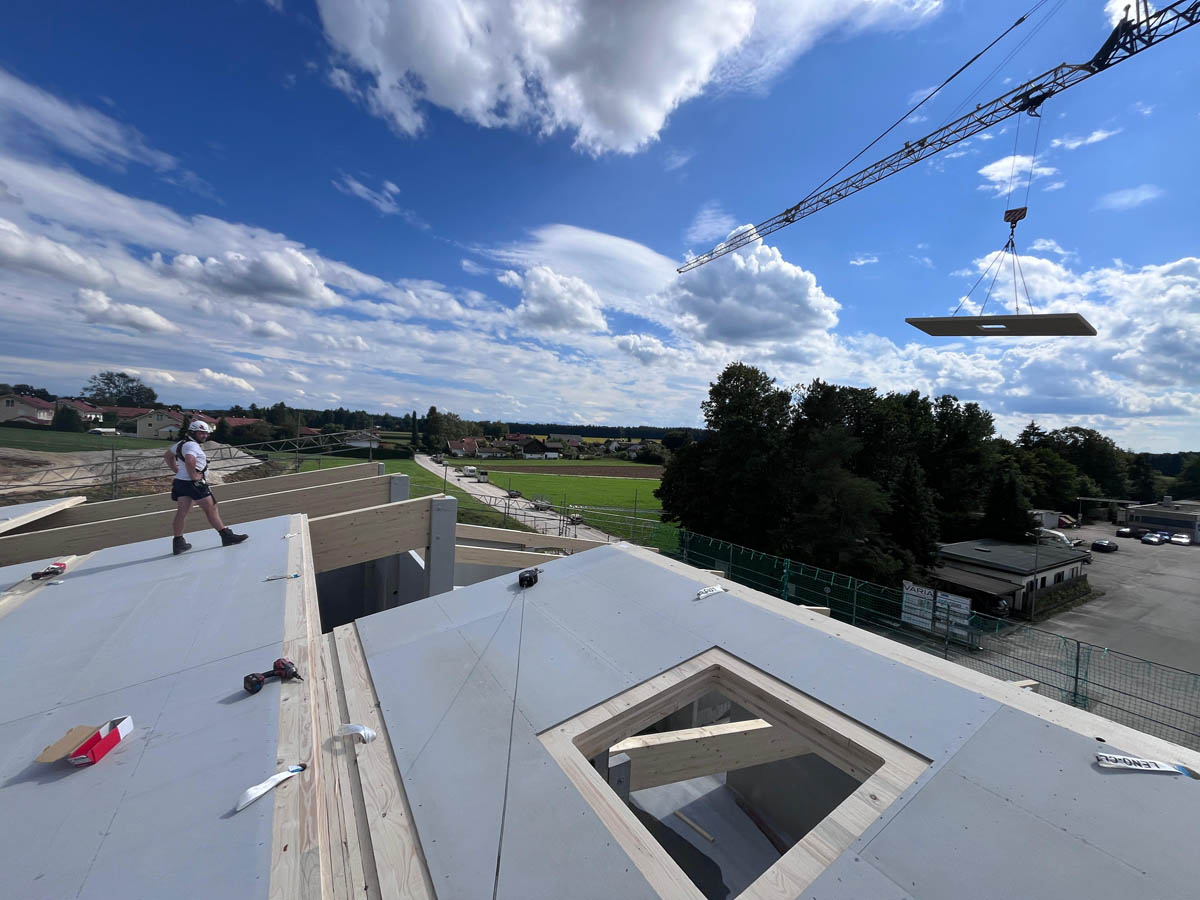 Bauarbeiter mit Helm steht auf einer Holzterrasse einer Baustelle, während ein großer Kran ein Bauteil anhängt, umgeben von Feldern und Gebäuden unter blauem Himmel mit Wolken.