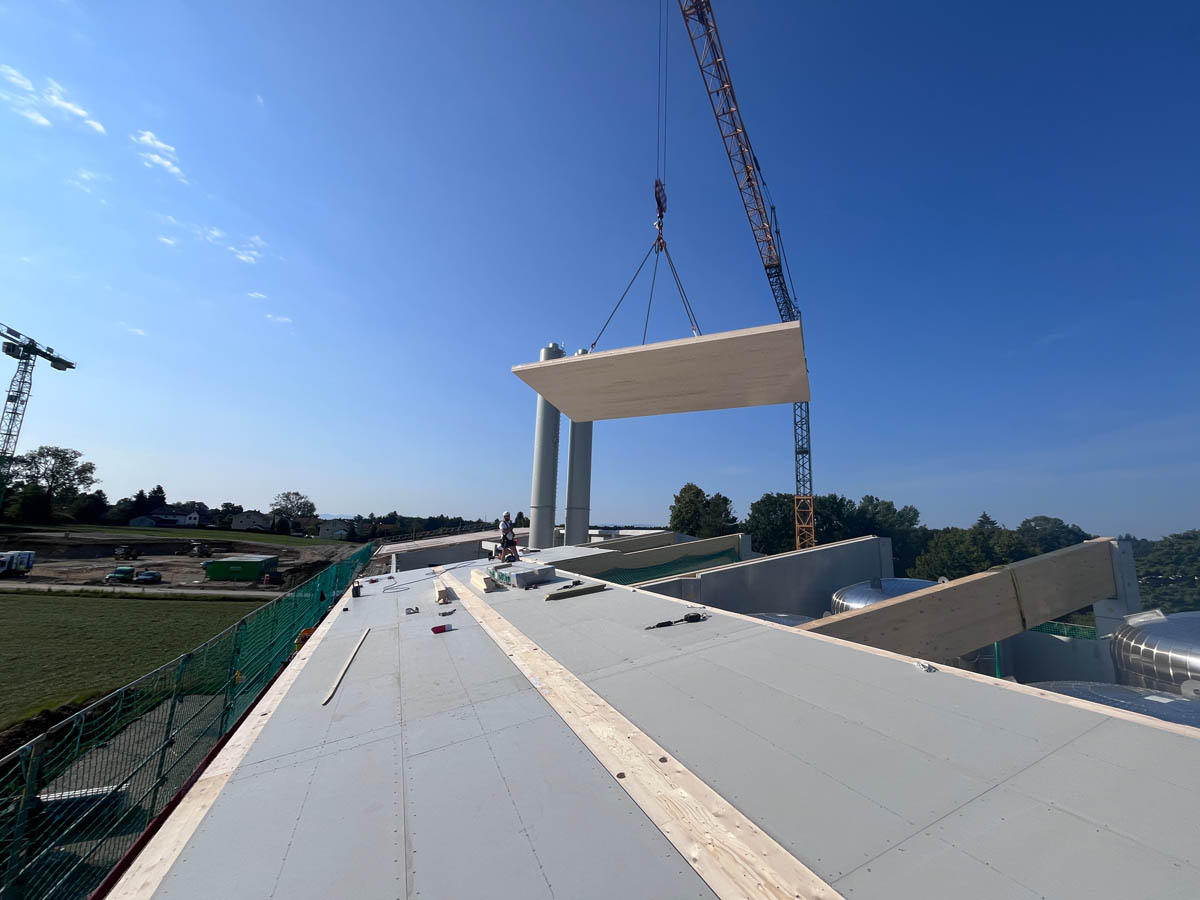Baustelle mit Kran, der ein großes Holzpaneel über einem flachen Dach hebt, unter blauem Himmel.