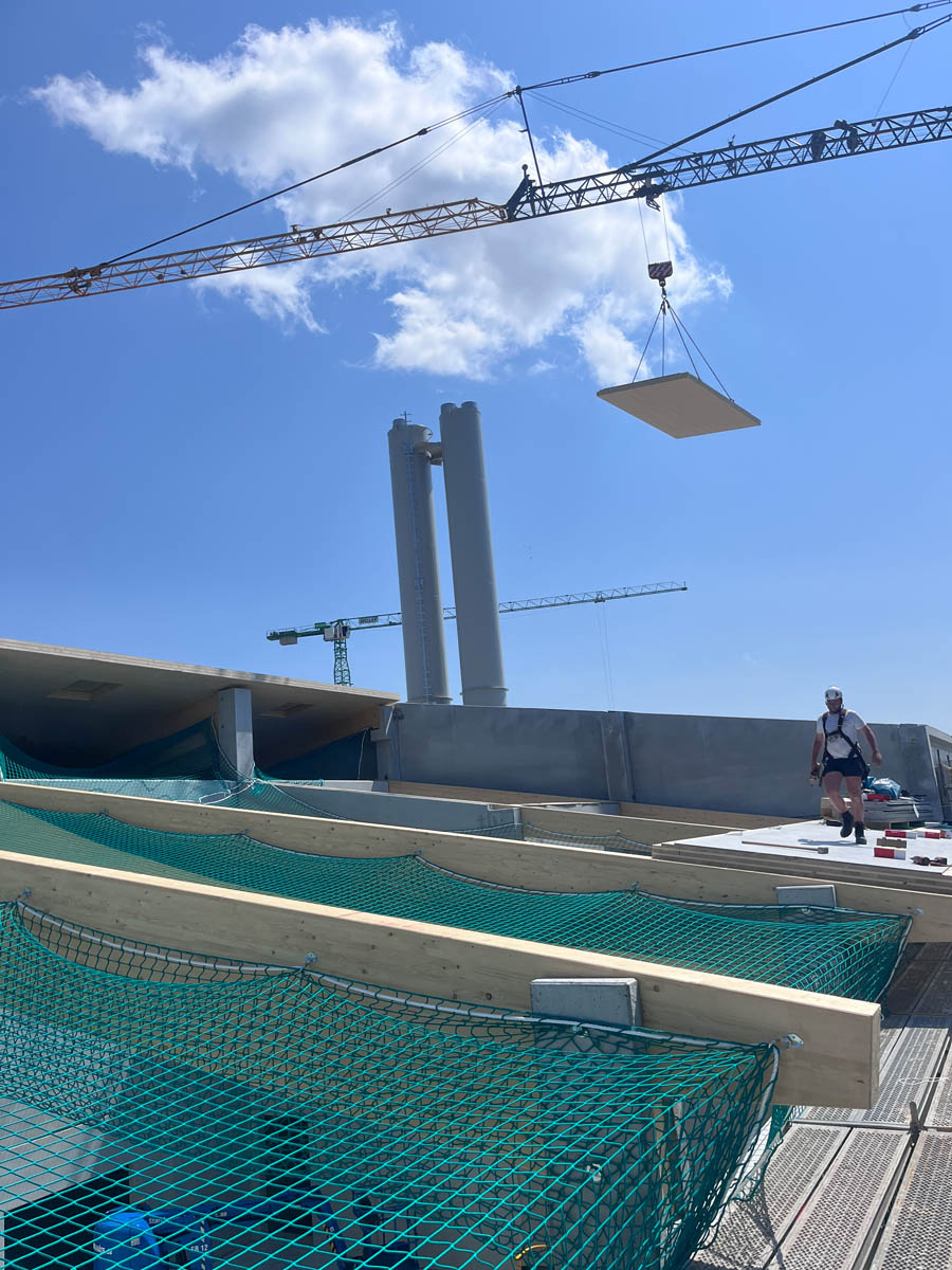 Baustelle mit Kränen, schwebendem Betonelement und Arbeiter auf einem Gebäude unter blauem Himmel.