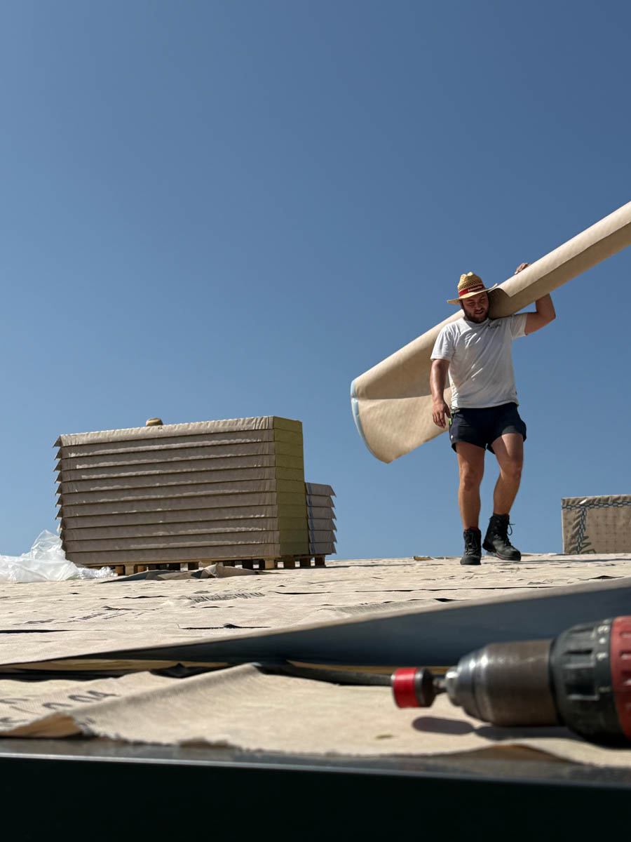 Ein Mann mit Strohhut trägt eine große Rolle Dachbahn auf einer Baustelle bei blauem Himmel.