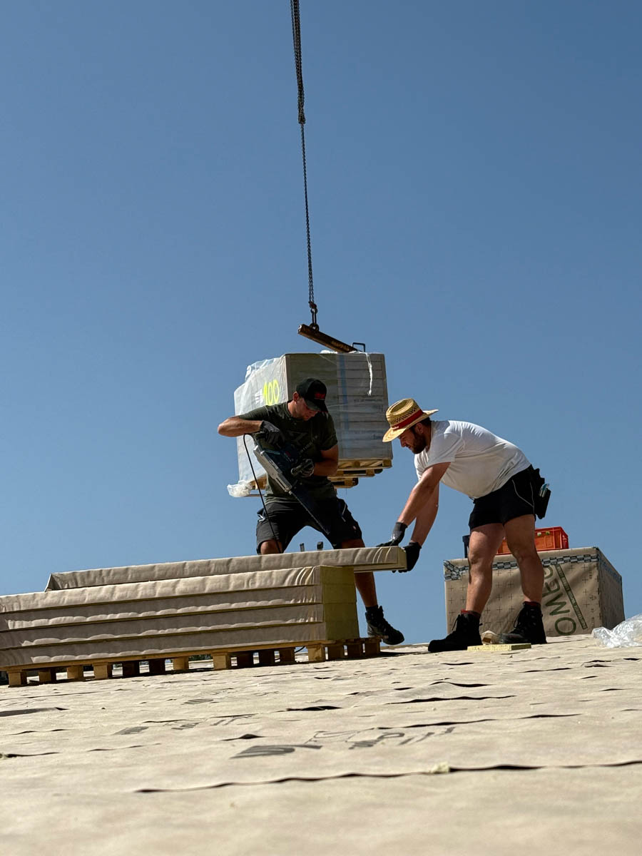Zwei Bauarbeiter heben gemeinsam eine Platte auf einer Baustelle unter klarem blauem Himmel.
