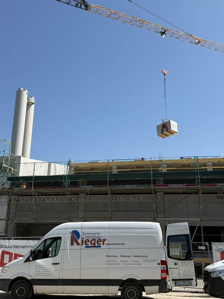 Weißer Handwerker-Van der Zimmerei Holzbau Rieger vor einer Baustelle mit Gerüst und Kran, der einen Anhänger mit Material hebt.