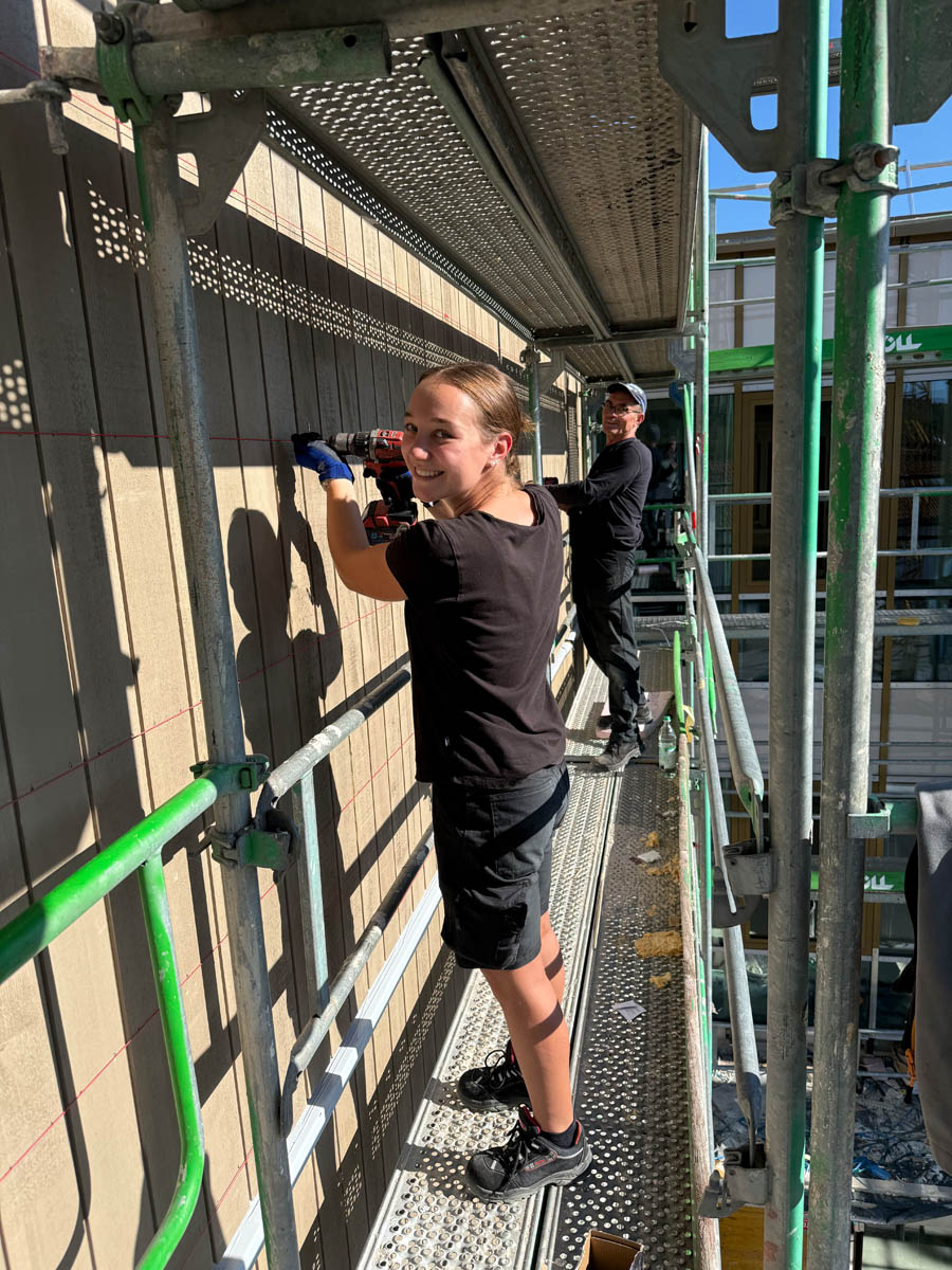 Zwei Bauarbeiter auf einem Baugerüst, die an einer Außenwand arbeiten, eine junge Frau bedient einen Bohrer.