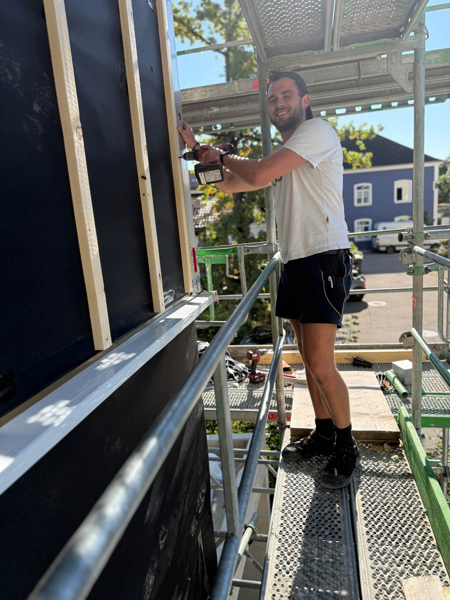 Ein Mann in weißem T-Shirt und schwarzer Hose arbeitet mit einem Akkuschrauber auf einem Gerüst an einem Außenwandrahmen.