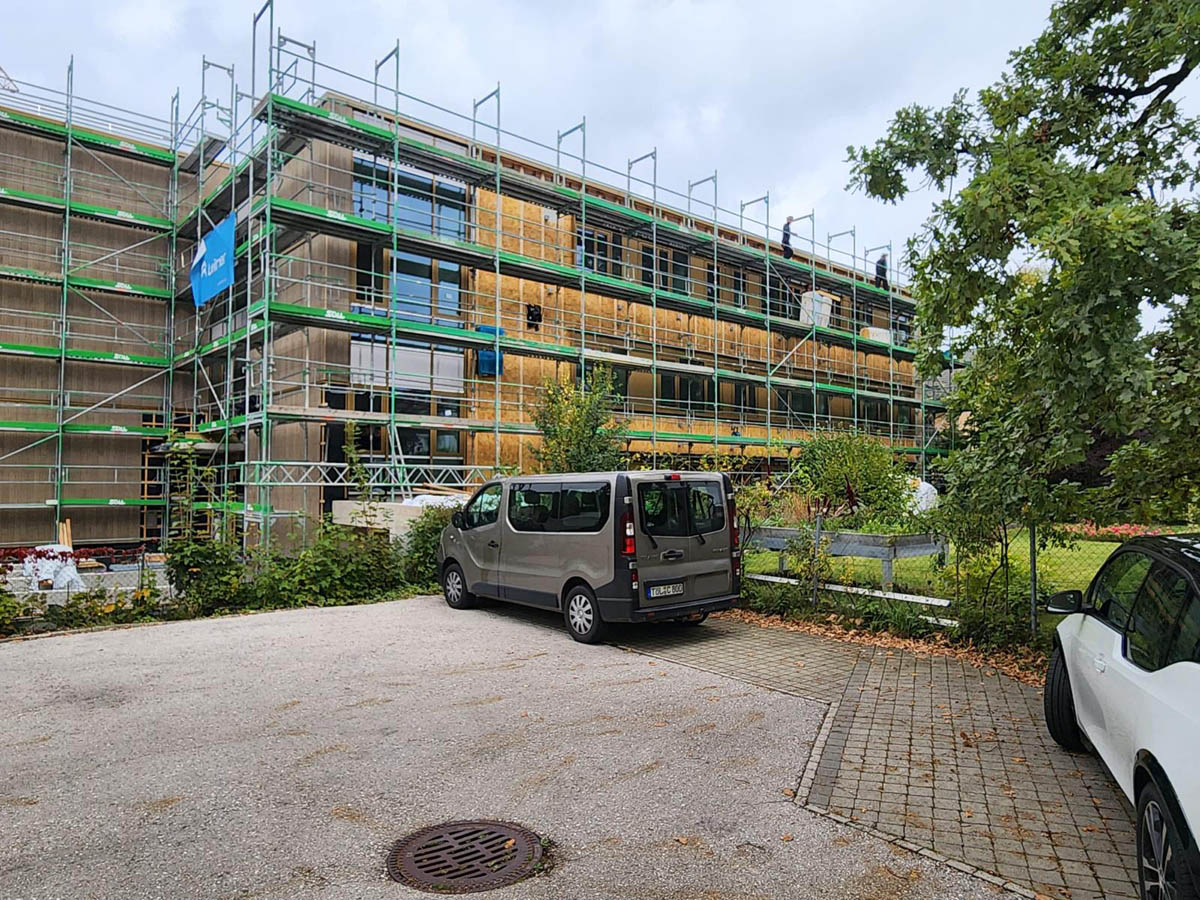 Gebäude unter Renovierung mit Gerüst, Parkplätze im Vordergrund und zwei geparkte Fahrzeuge.