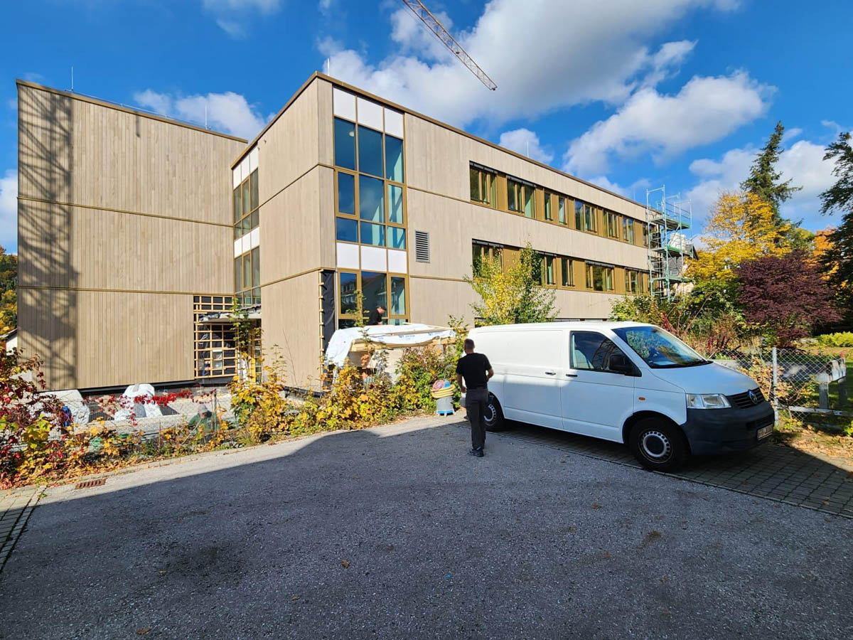 Modernes dreistöckiges Gebäude mit Holzfassade, einem weißen Lieferwagen und einer Person auf dem Parkplatz bei sonnigem Himmel.