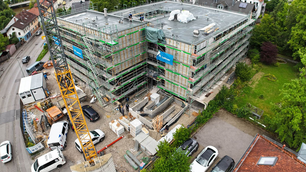 Baustelle eines mehrstöckigen Gebäudes mit Gerüst und Kran, mehreren Fahrzeugen und Bauarbeitern auf einem Grundstück neben einer Straße und einem grünen Garten.