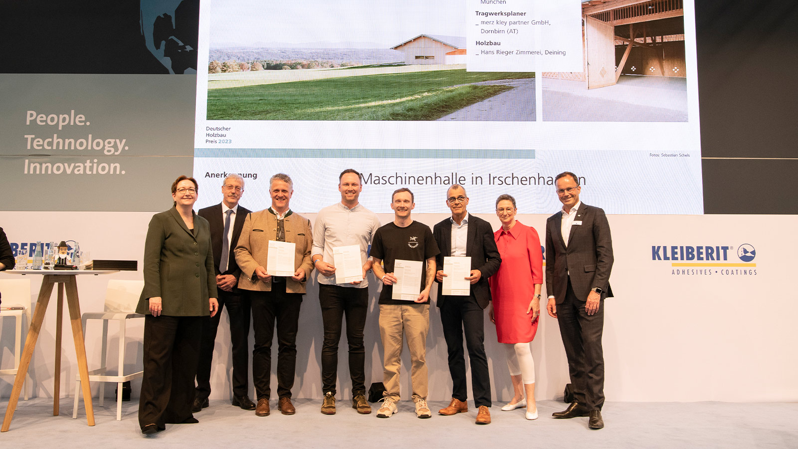 Gruppenfoto bei der Preisverleihung des Deutschen Holzbaupreises 2023 mit den Beteiligten der Maschinenhalle in Irschenhausen auf einer Bühne vor einer Projektpräsentation.