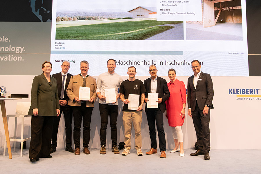 Gruppenfoto bei der Preisverleihung des Deutschen Holzbaupreises 2023 mit den Beteiligten der Maschinenhalle in Irschenhausen auf einer Bühne vor einer Projektpräsentation.