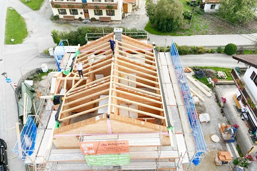 Luftaufnahme eines im Bau befindlichen Hauses mit sichtbarem Holzrahmen und Dachkonstruktion, umgeben von Gerüsten und Baumaterialien, mit Arbeitern auf dem Dach, in einem Wohngebiet.