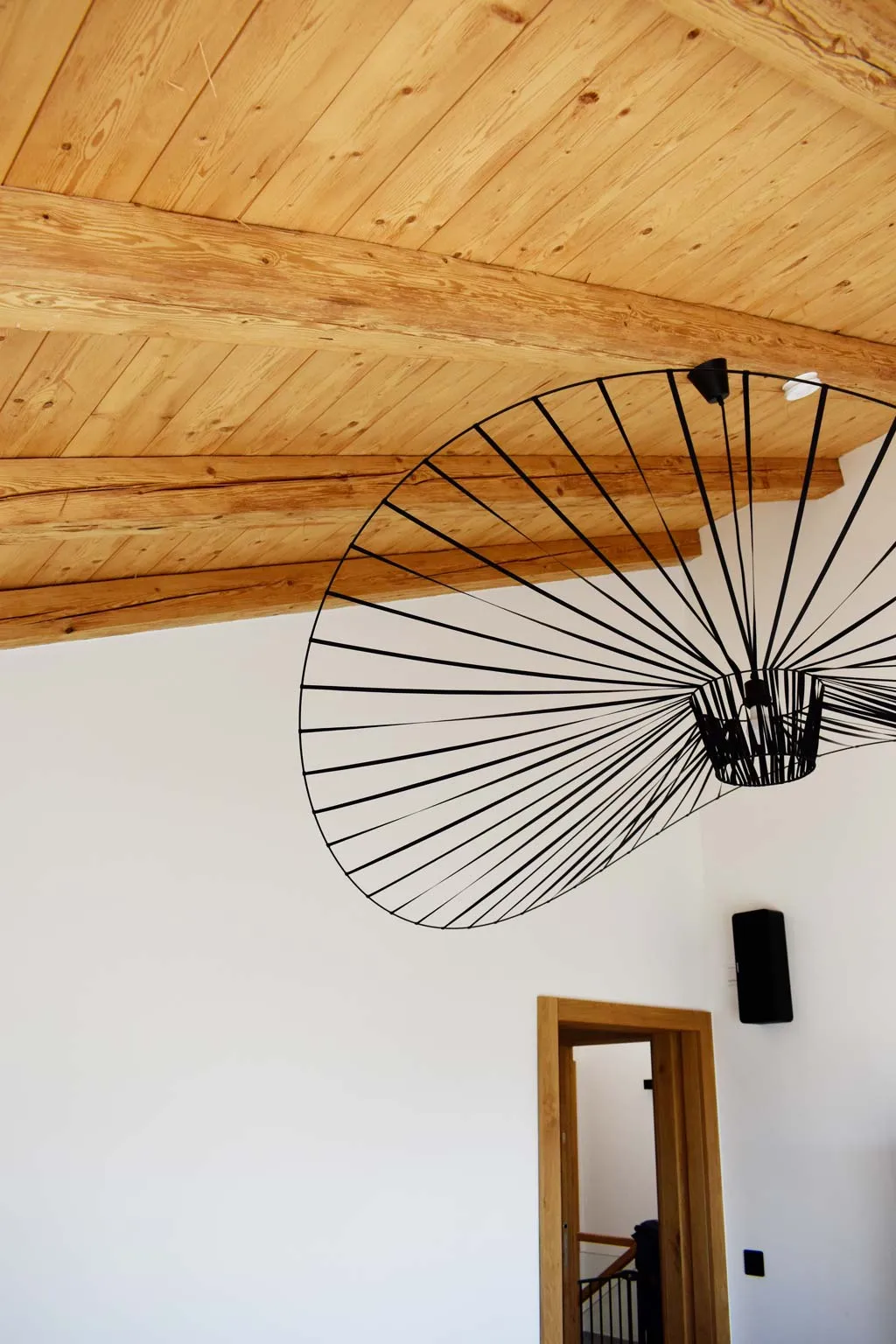 Innenansicht eines Raumes mit einem hölzernen Dachstuhl und einer modernen, schwarzen, spiralförmigen Hängeleuchte, darunter eine offene Tür mit Blick auf eine Treppe.