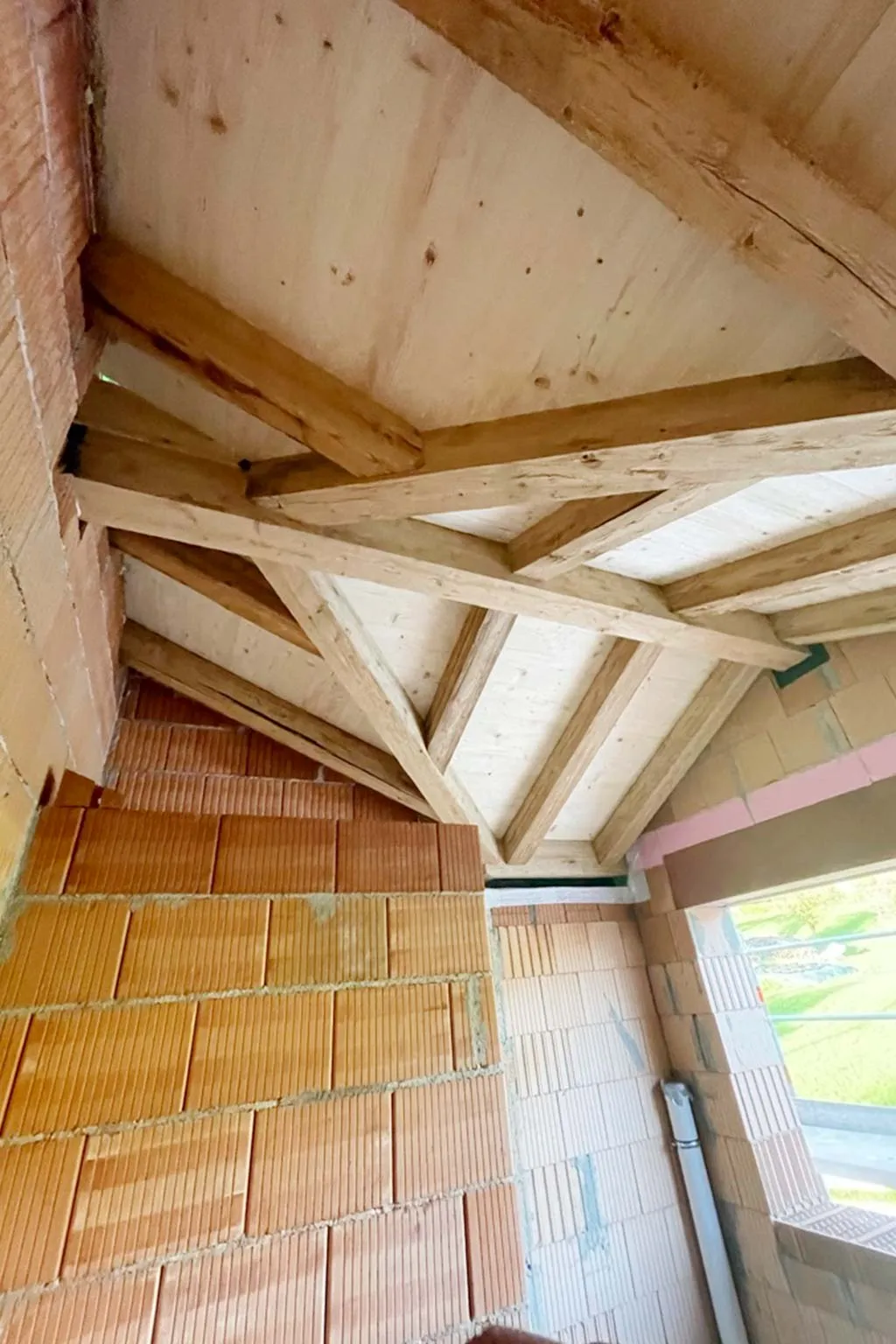 Innenansicht eines im Bau befindlichen Raumes mit einer Dachkonstruktion aus Holzbalken und unverputzten Ziegelwänden.