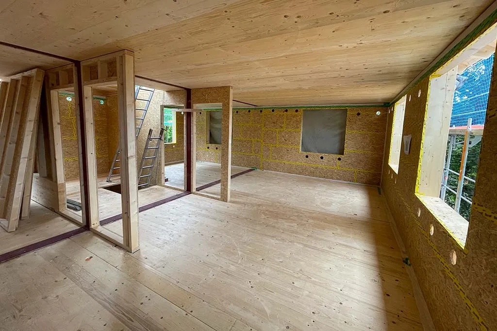 Innenansicht eines im Bau befindlichen Hauses mit sichtbaren Holzrahmen, unvollständigen Wänden und einer Leiter, die zu einem oberen Stockwerk führt.