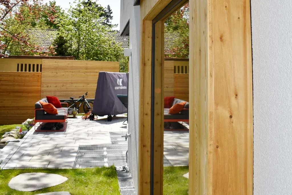 Blick auf eine Terrasse mit gepolsterten Gartenmöbeln und einem abgedeckten Tischtennistisch, umgeben von einem Holzzaun und grünen Bäumen, aufgenommen durch eine offene Tür.