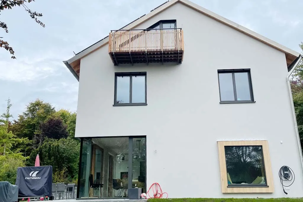 Rückansicht eines modernen Hauses mit einem Holzbalkon, großen Fenstern und einer Terrasse, auf der Gartenmöbel und ein abgedeckter Tischtennistisch stehen, umgeben von grüner Vegetation.