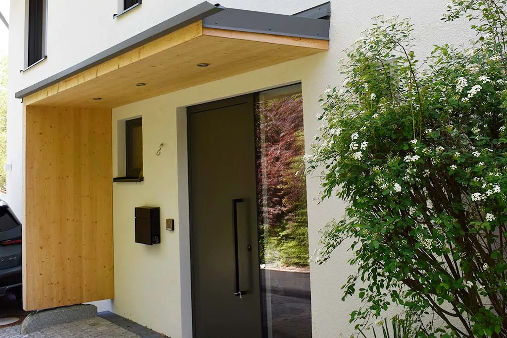 Eingangsbereich eines modernen Hauses mit einer grauen Tür und einem hölzernen Vordach, daneben ein blühender Strauch mit weißen Blumen.