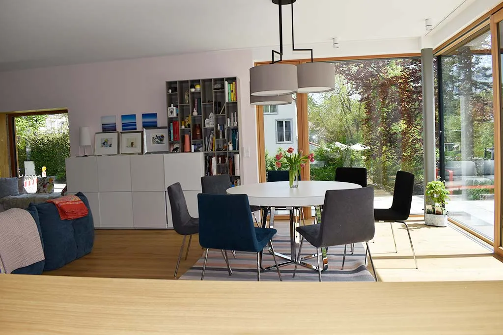 Helles und modernes Wohnzimmer mit großen Fenstern, einem runden Esstisch mit schwarzen Stühlen, einem Bücherregal und einem Sofa mit bunten Kissen.
