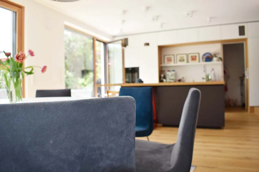 Moderner Wohn- und Essbereich mit einem runden Esstisch, dunkelblauen Stühlen, einem Blumenstrauß auf dem Tisch und einer offenen Küche im Hintergrund.
