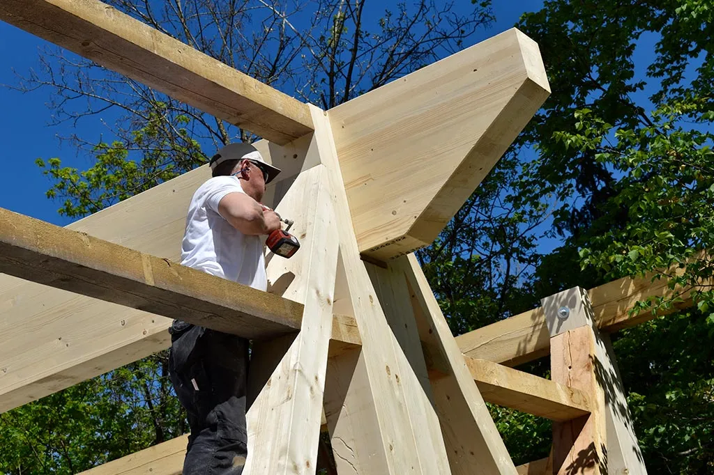 Ein Bauarbeiter in weißem T-Shirt und Schutzhelm befestigt Holzrahmen mit einer Bohrmaschine an einer Holzkonstruktion, bei sonnigem Wetter und blauem Himmel.