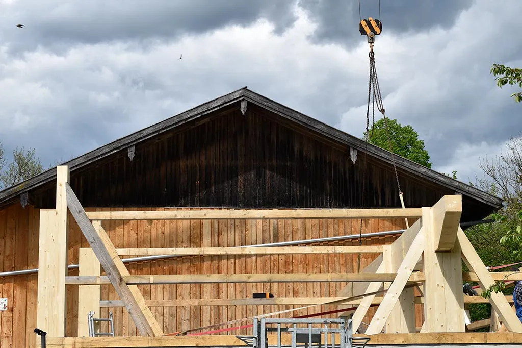 Teilweise fertiggestellte Holzkonstruktion vor einer alten Scheune, ein Kranhaken hängt darüber, bei bewölktem Himmel.