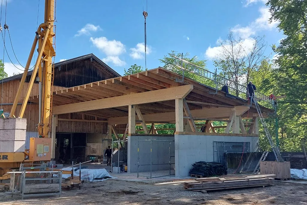 Bau einer landwirtschaftlichen Scheune mit hölzernem Dachrahmen und Betonfundament, Arbeiter auf dem Dach und ein gelber Kran im Vordergrund, bei sonnigem Wetter.