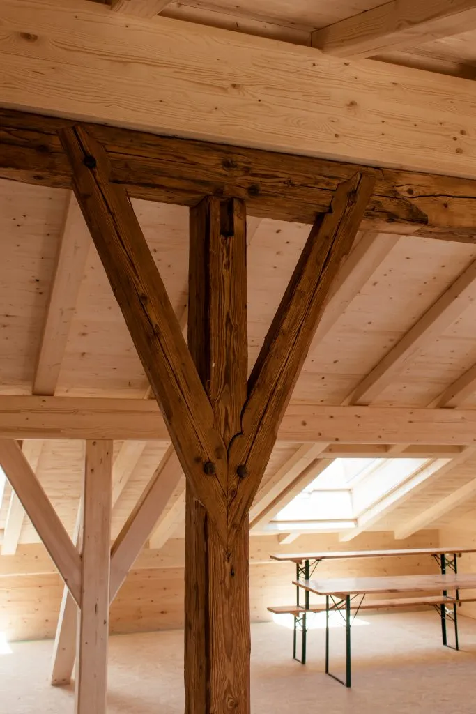 Innenansicht eines Dachbodens mit sichtbaren Holzbalken, einer rustikalen Holzstütze und einem Tisch mit Bänken unter einem Dachfenster.